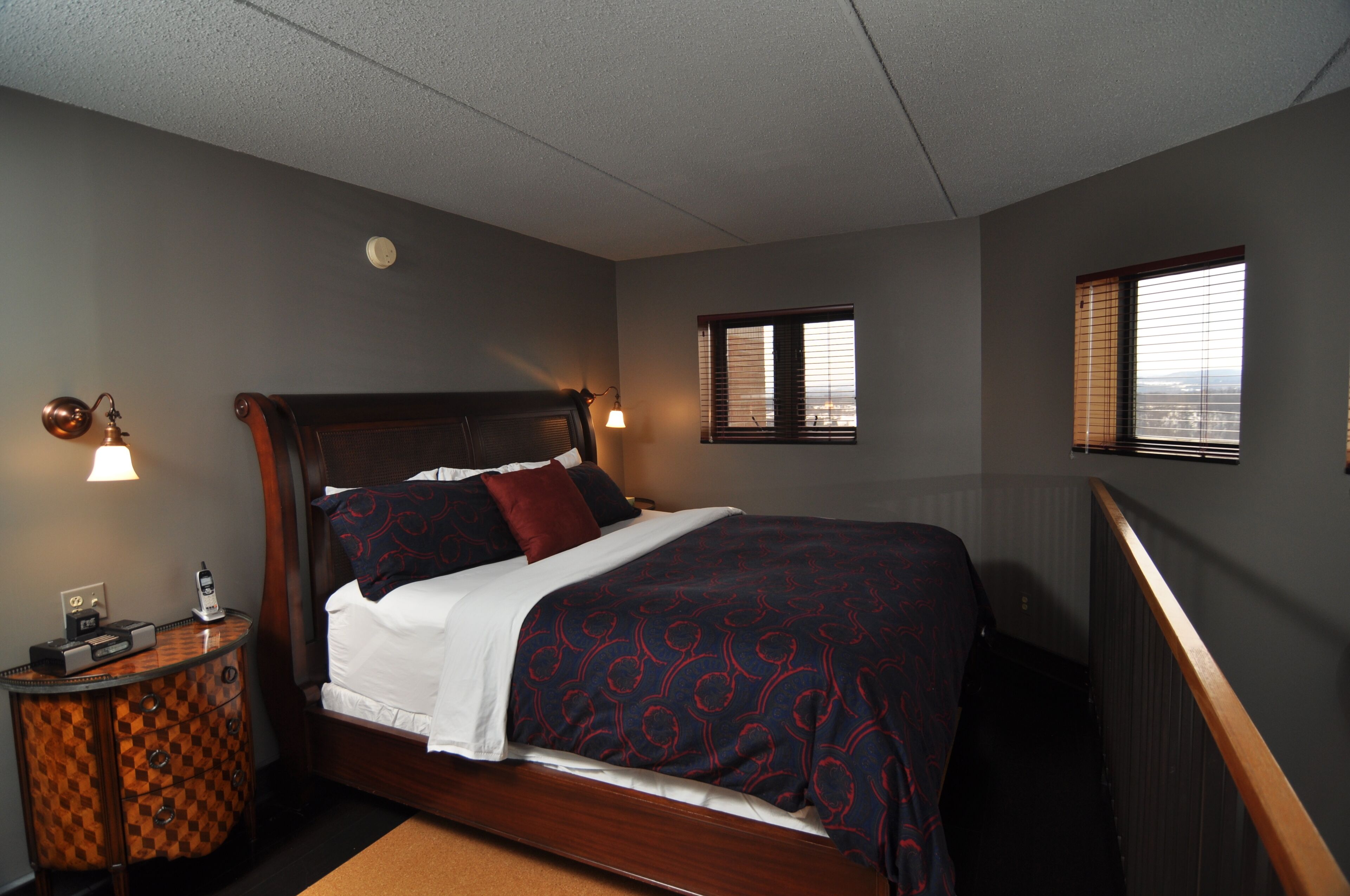 A hotel room with a wooden bed, two bedside lamps, a nightstand, and a window with blinds.