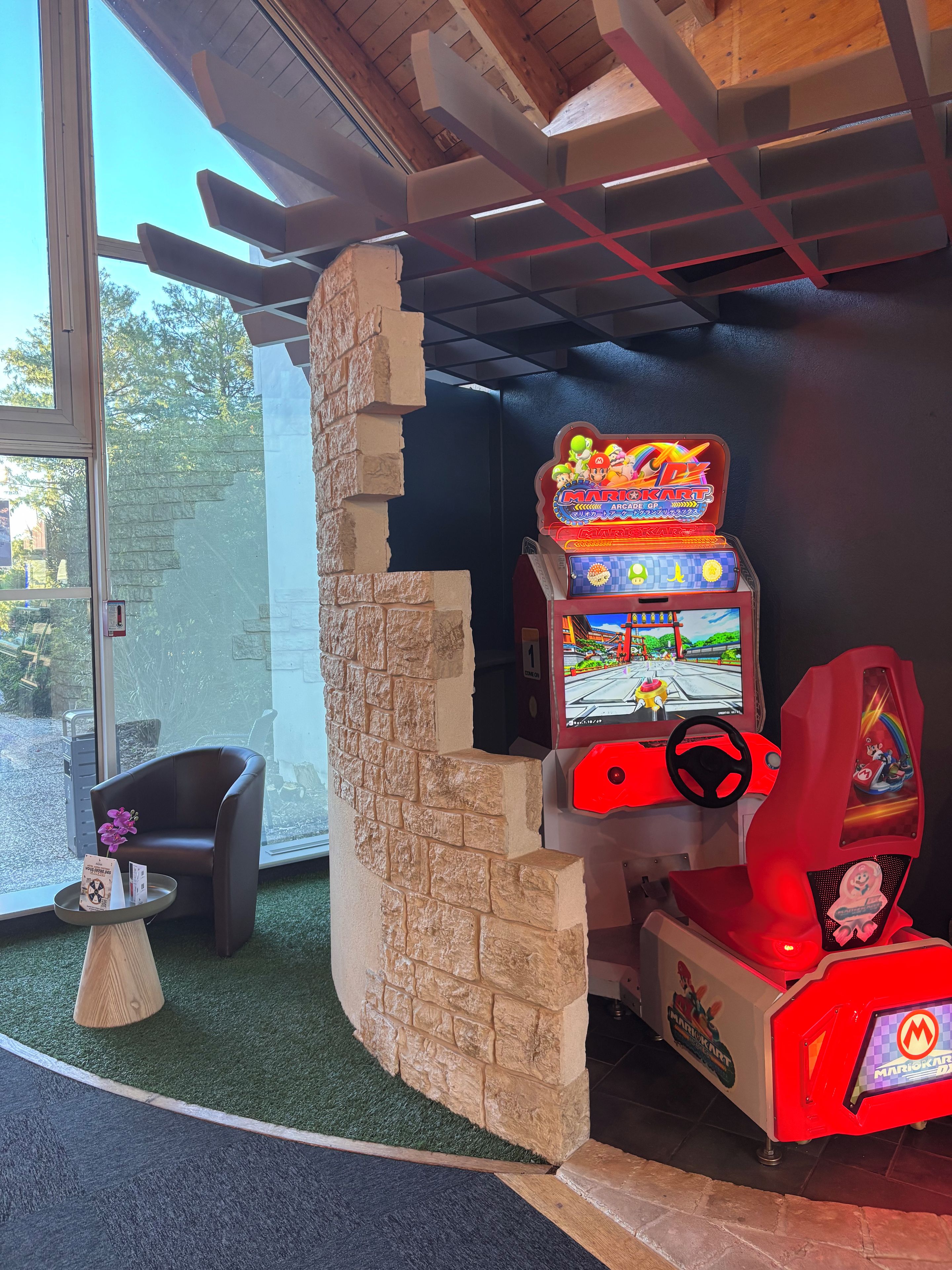 Een Mario Kart-automaat met een stenen muur als achtergrond, een gezellige zithoek met een klein tafeltje en uitzicht door een groot raam.