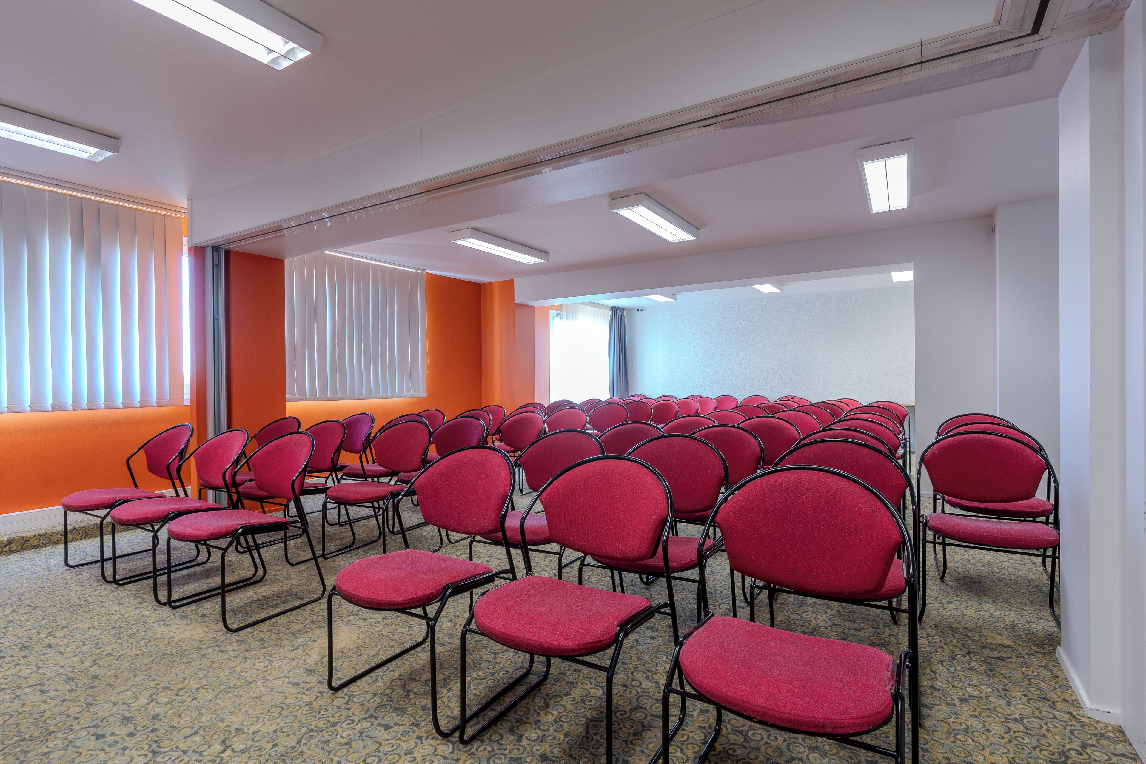 Una sala de conferències amb files de cadires vermelles, parets blanques i detalls taronja.
