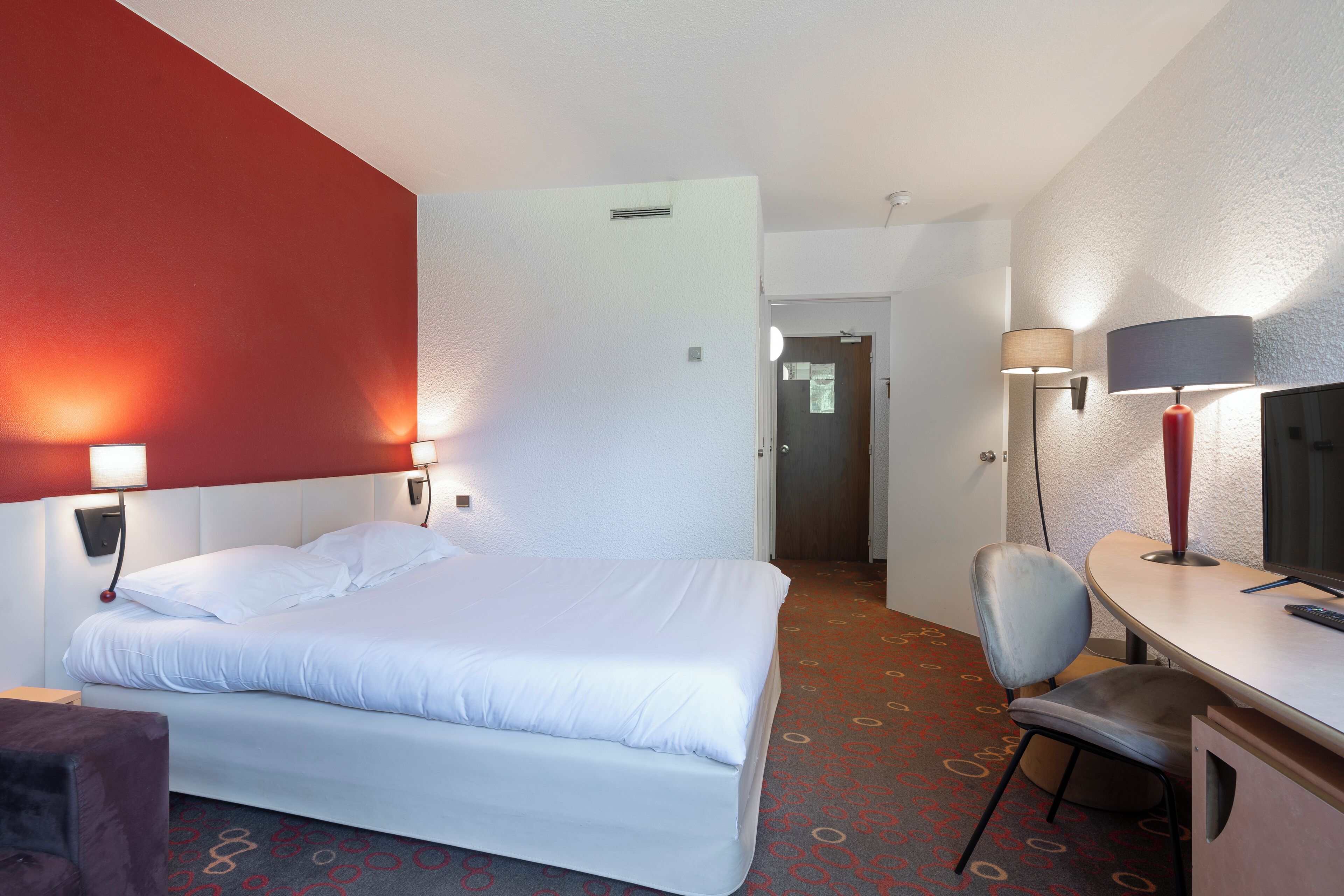 Quarto de hotel com cama, escrivaninha e cadeira. Uma porta dá para outro cômodo.