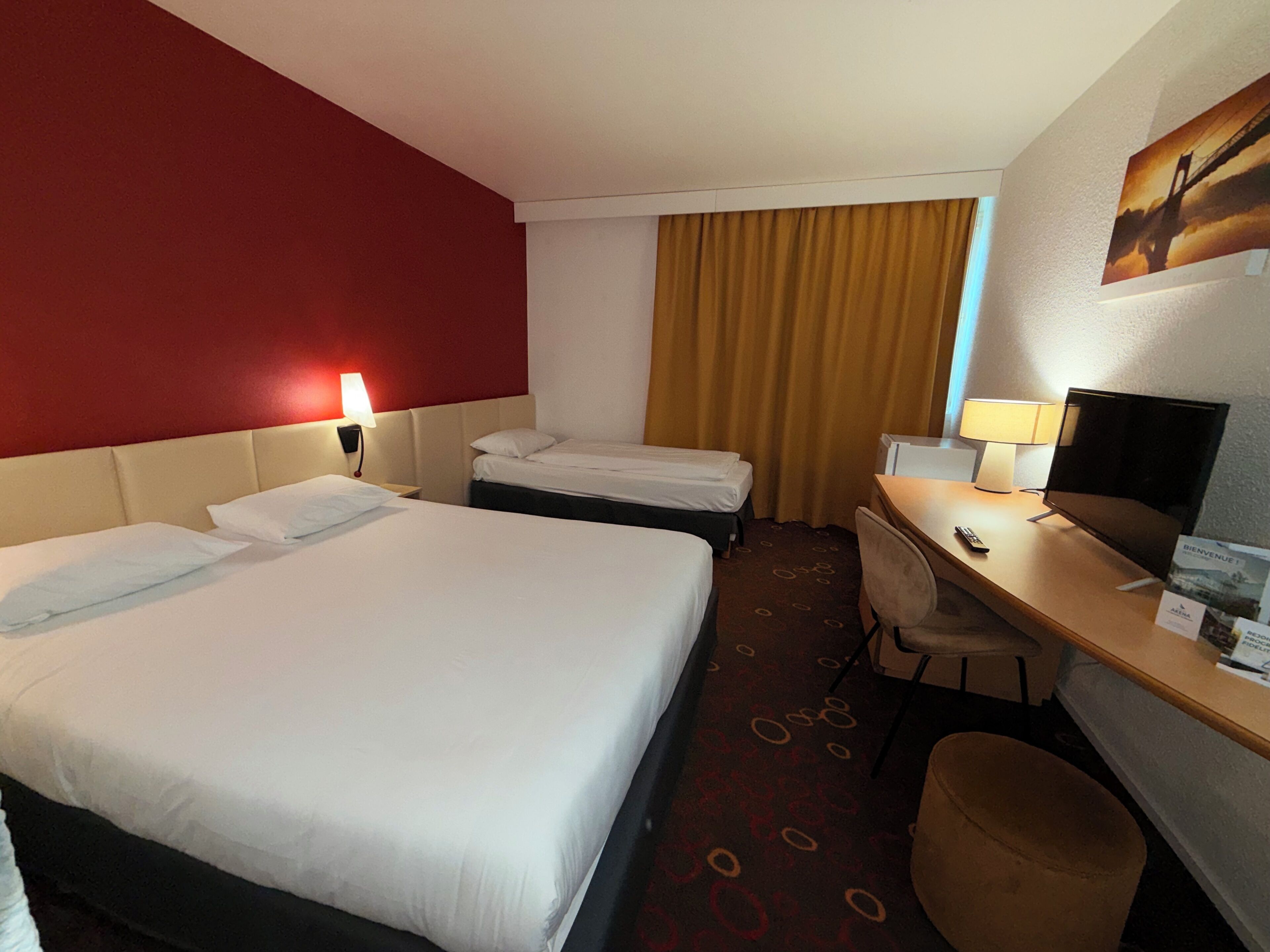 A hotel room with two beds, a desk, a chair, a TV, and a red accent wall.