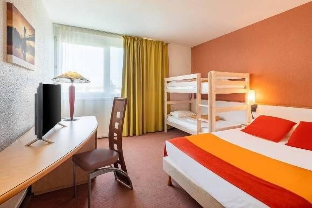 A hotel room with bunk beds, a desk with a lamp, a chair, and a bed with red pillows.