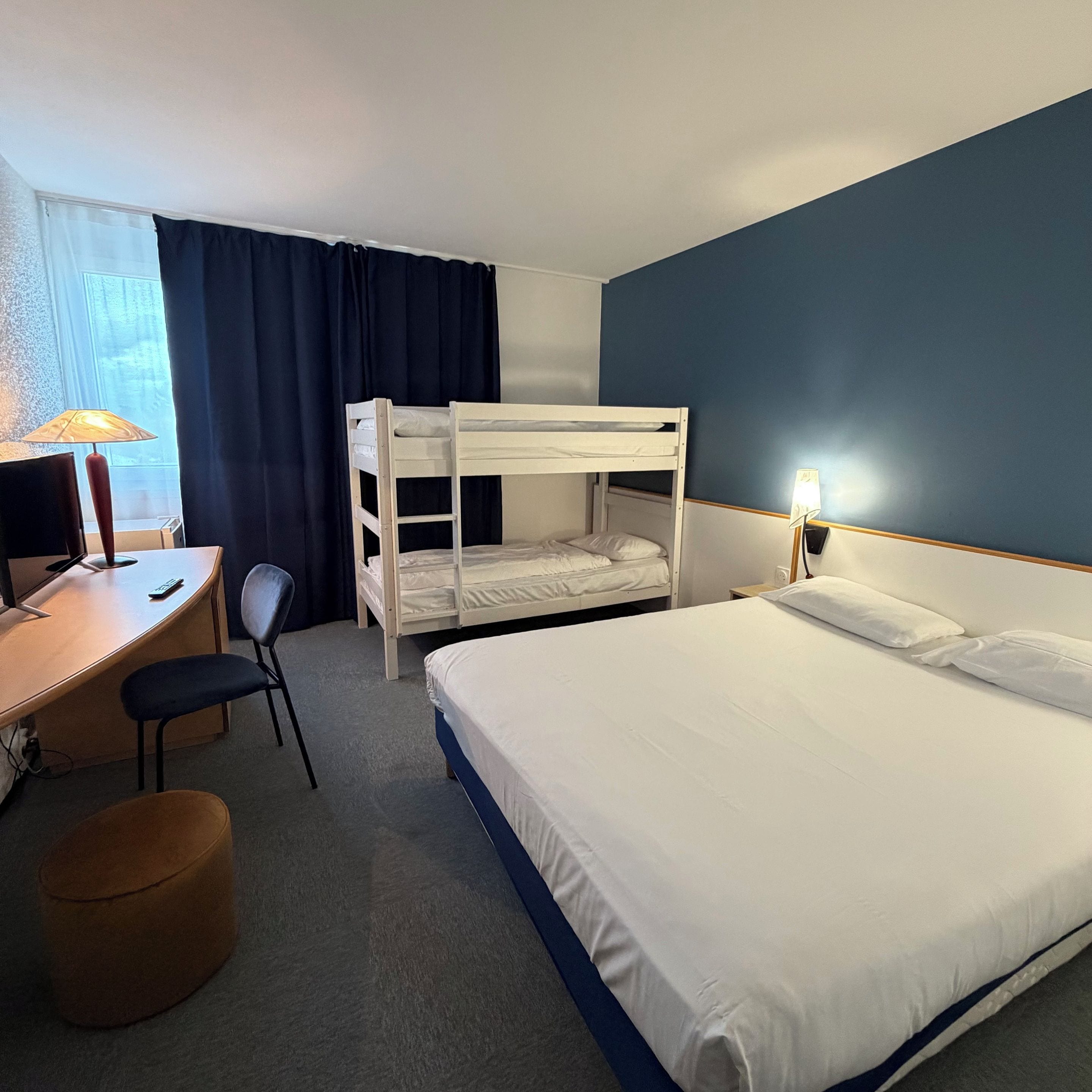 A hotel room with a bunk bed, a desk with a lamp, a chair, and a bedside table with a lamp.