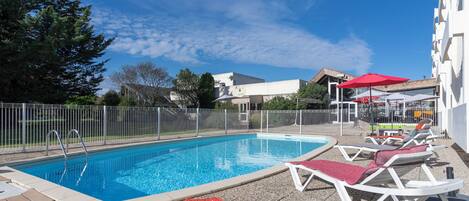 A swimming pool with lounge chairs and umbrellas, surrounded by a fence and buildings.