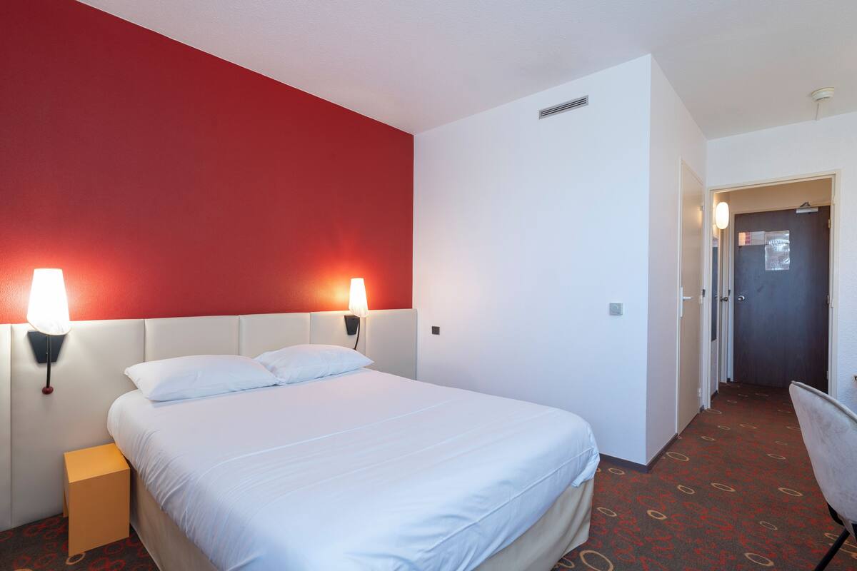 A hotel room with a bed, two bedside lamps, a nightstand, a chair, and a door leading to another room.
