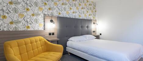 Een hotelkamer met een bed, een gele fauteuil, een hoofdeinde met wandlampen en een behang met een bloemenprint.