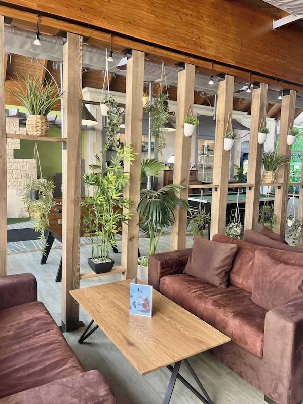 A cozy seating area with a wooden table, cushioned sofas, and hanging plants. - HĂŽtel AKENA POITIERS NORD FUTUROSCOPE (Chasseneuil-du-Poitou)