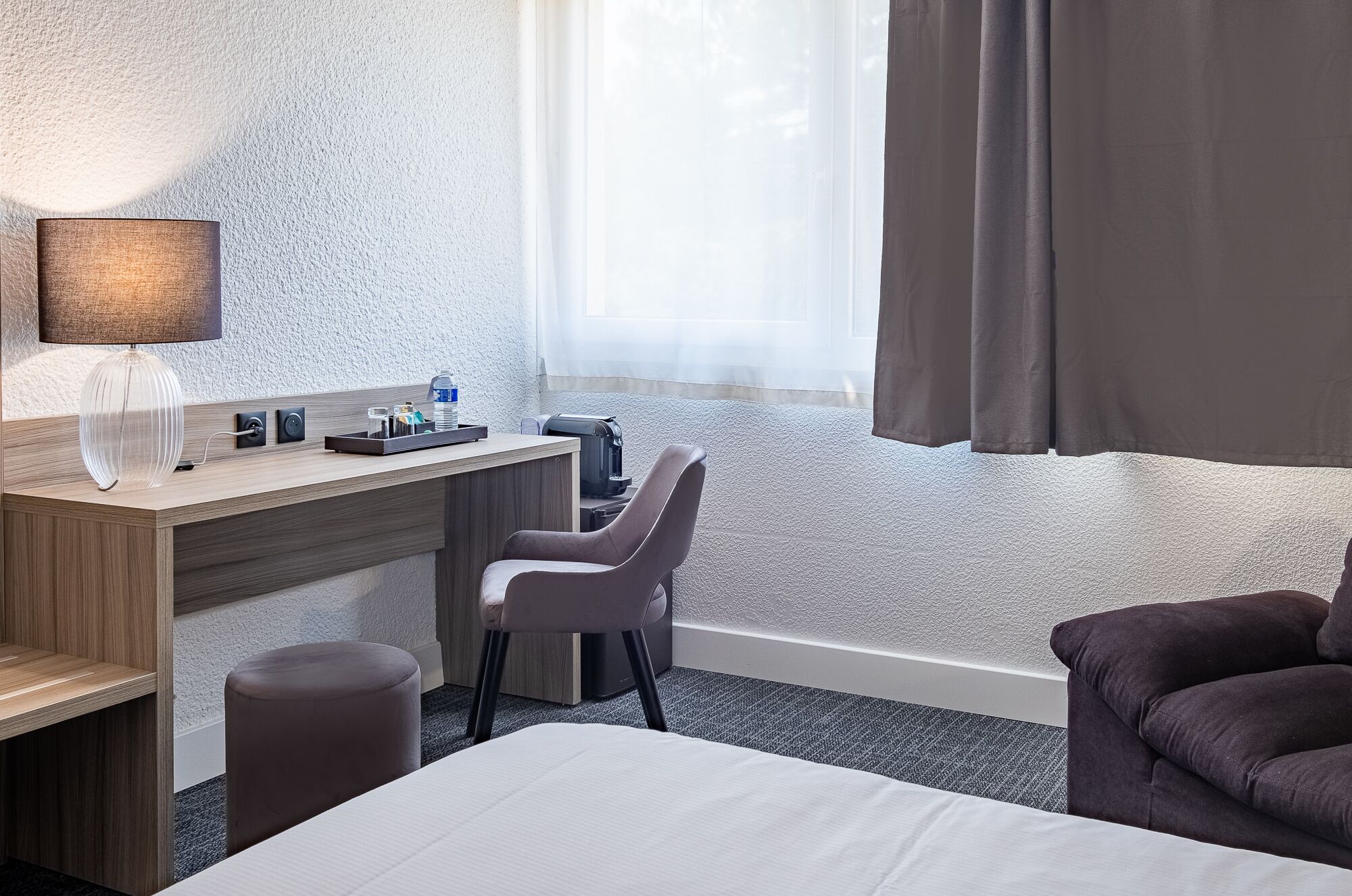 A hotel room with a bed, a desk with a lamp, a chair, a coffee machine, and a window with curtains.