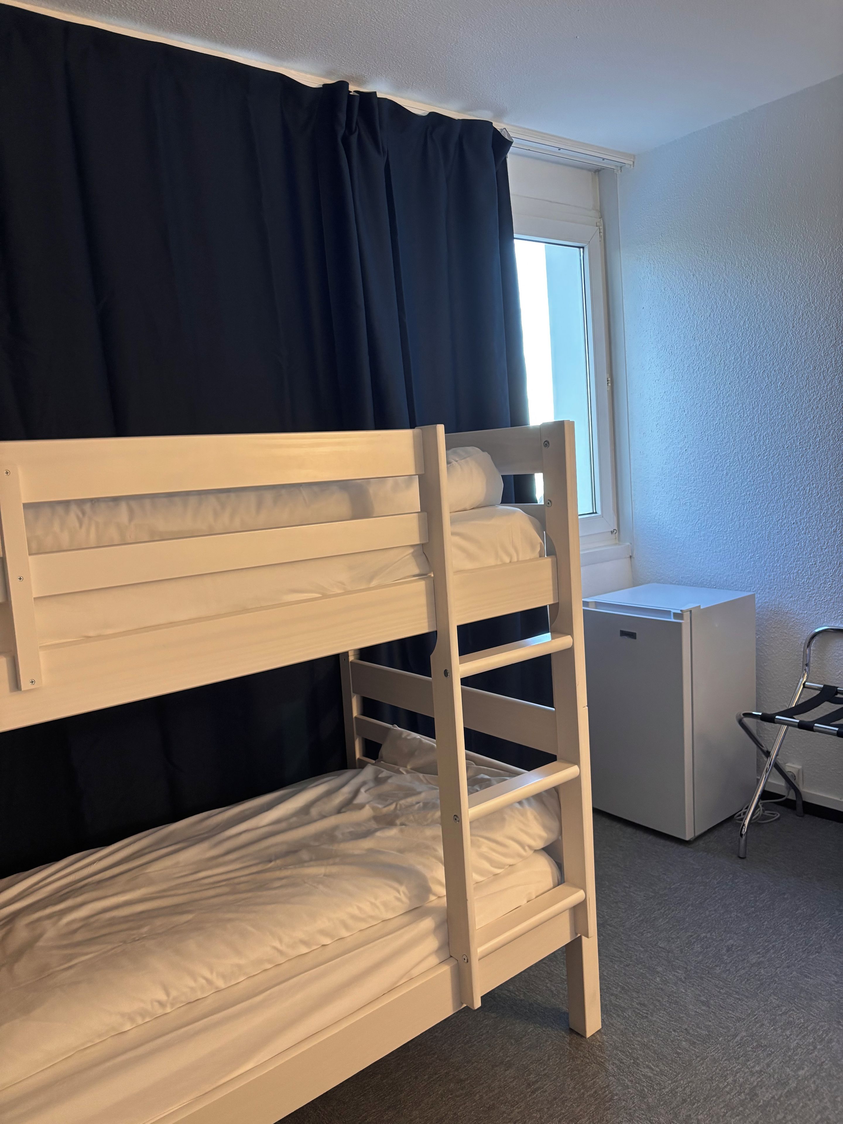 En køjeseng med hvid ramme, et lille skrivebord og en stol i et rum med et vindue og mørke gardiner.