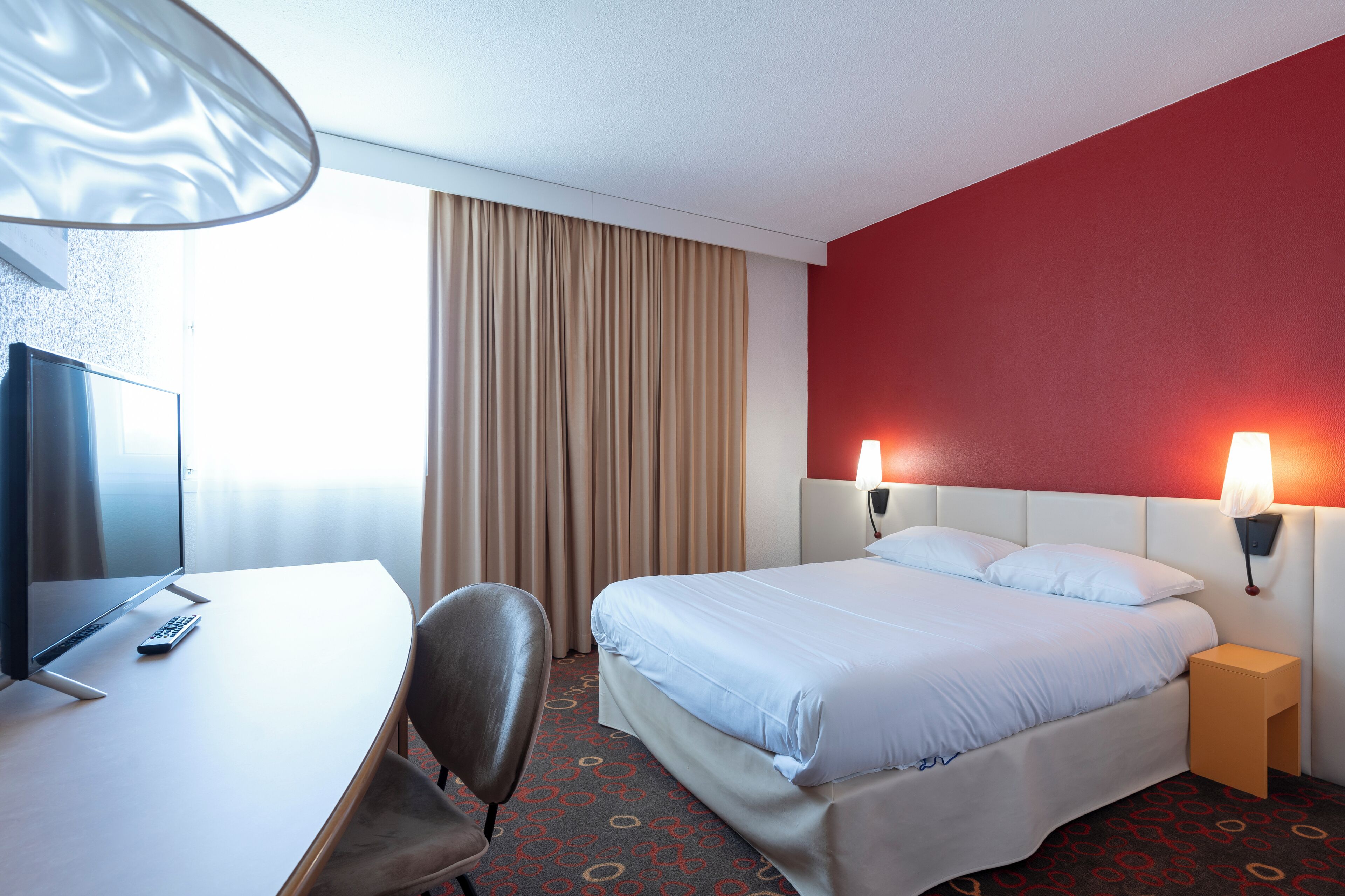 Habitación de hotel con cama, mesita de noche, lámpara, escritorio, silla, televisor y cortinas.