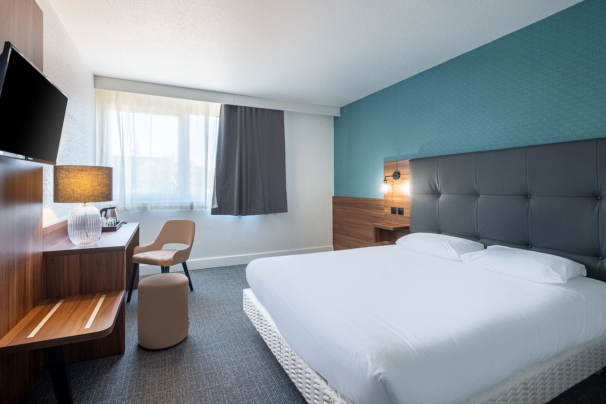 A hotel room with a bed, a desk with a lamp, a chair, a TV, and a window with curtains.