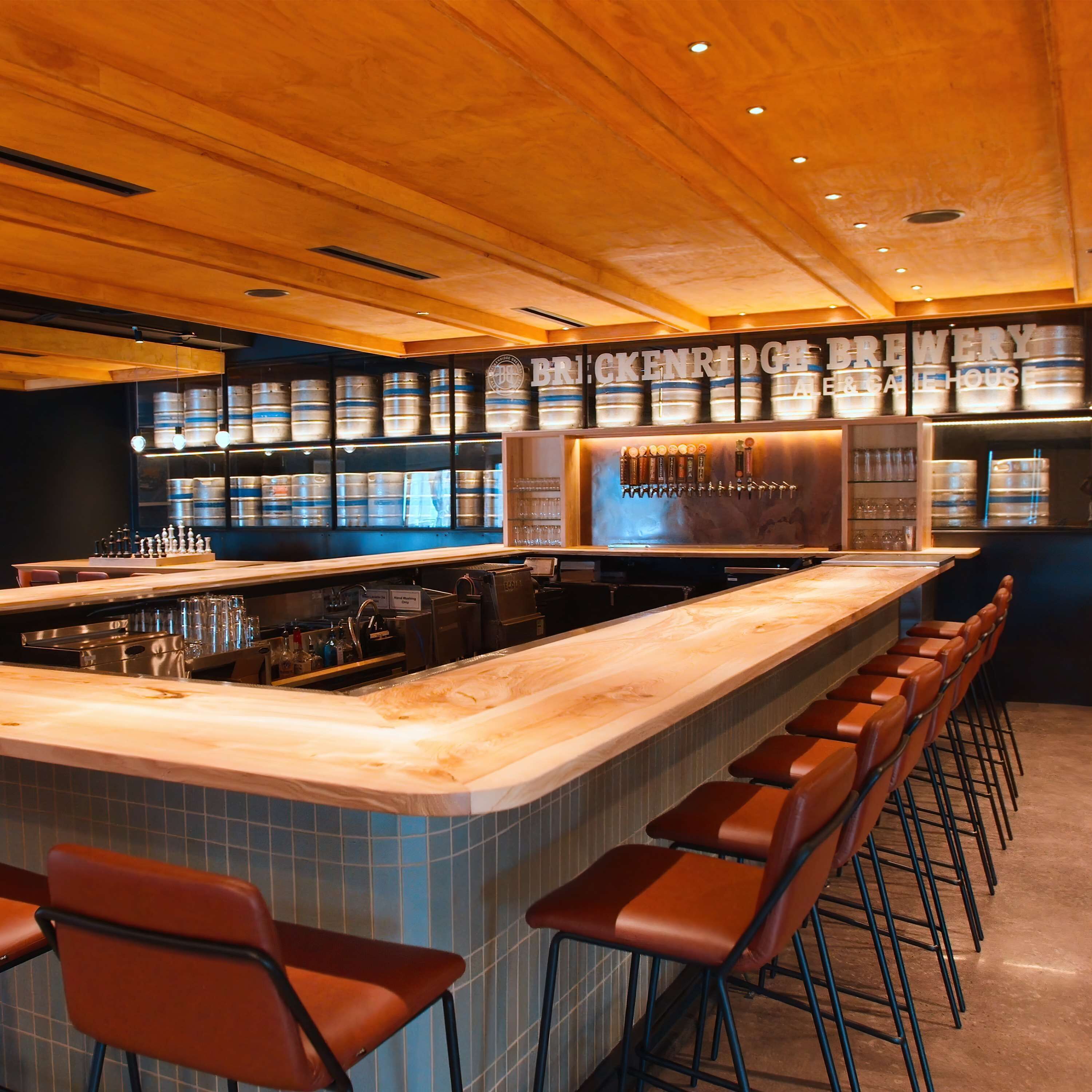 Zona de bar con una barra alargada, varios taburetes y una pared que muestra barriles de cerveza y grifos.