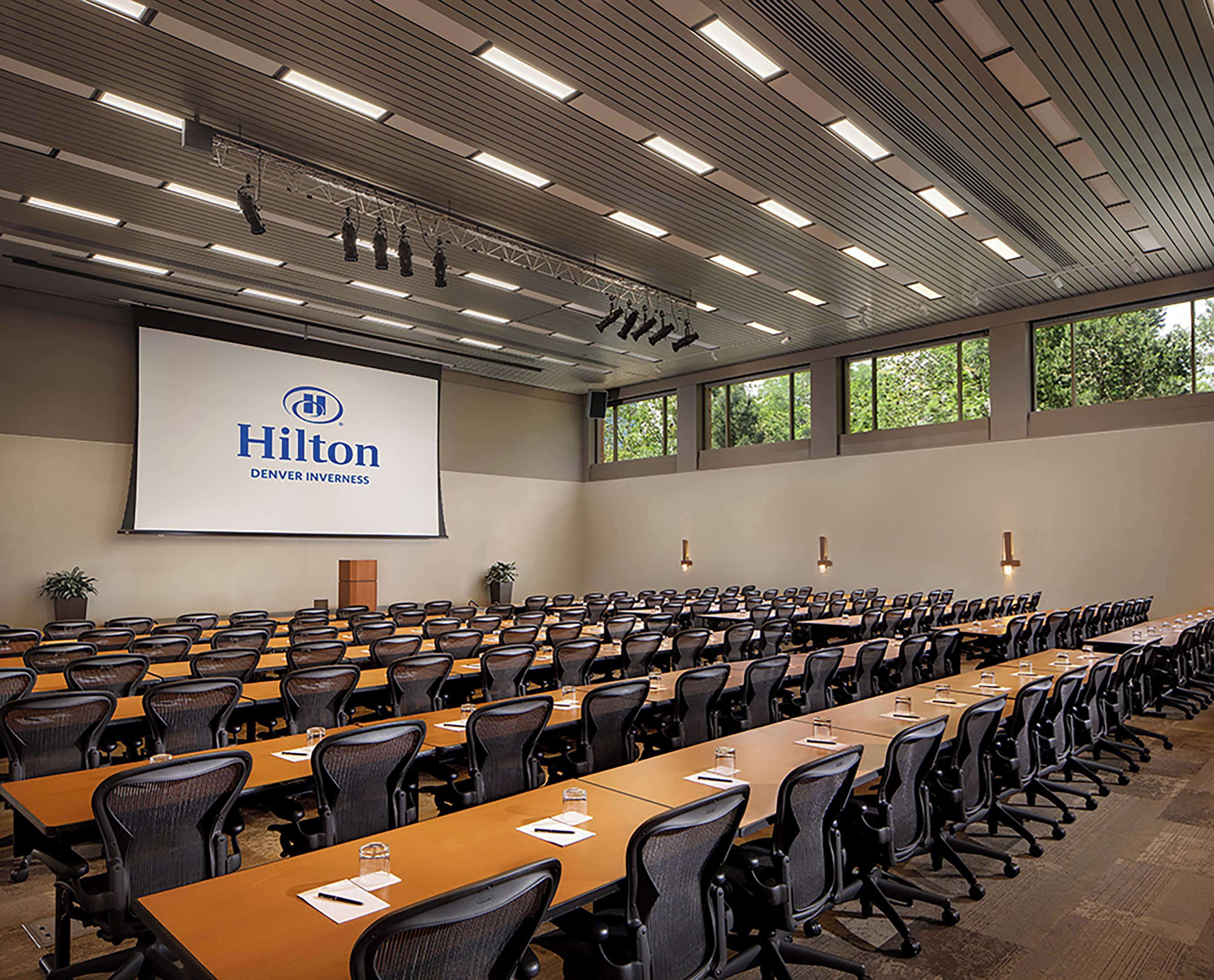 A large conference room with rows of tables and chairs, a projection screen displaying 'Hilton Denver Inness', and large windows with a view of greenery.