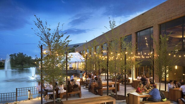 An outdoor seating area with wooden benches and tables, a brick building, and a fountain in the background.