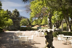 An outdoor seating area with white tables and chairs, surrounded by trees and a statue.