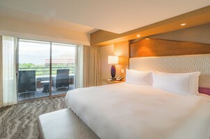 Habitación de hotel con una cama grande, una mesita de noche con lámpara, vistas al exterior por una puerta corredera y un balcón con sillas.