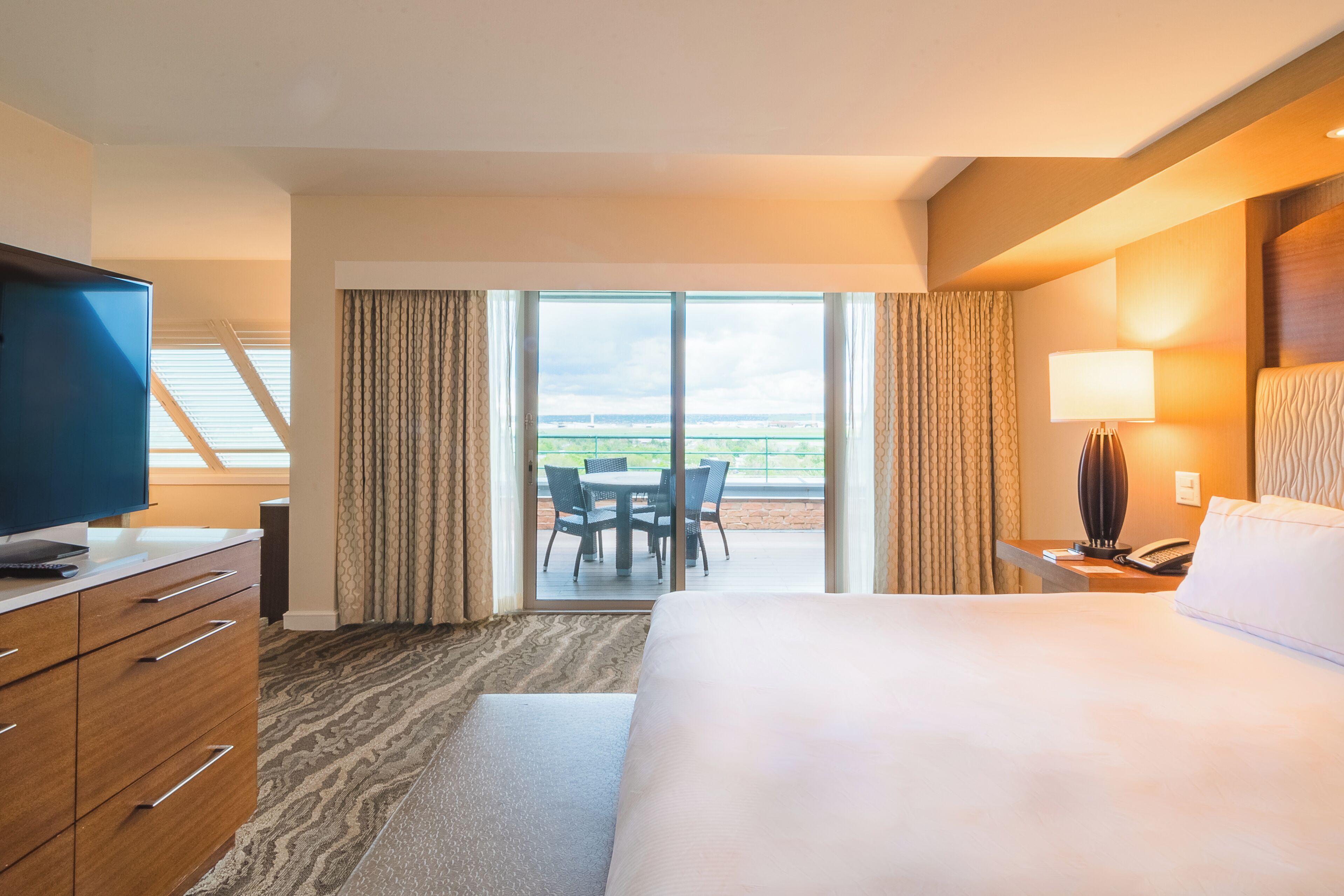 A hotel room with a large bed, a TV, a desk with a lamp, and a balcony with a table and chairs.