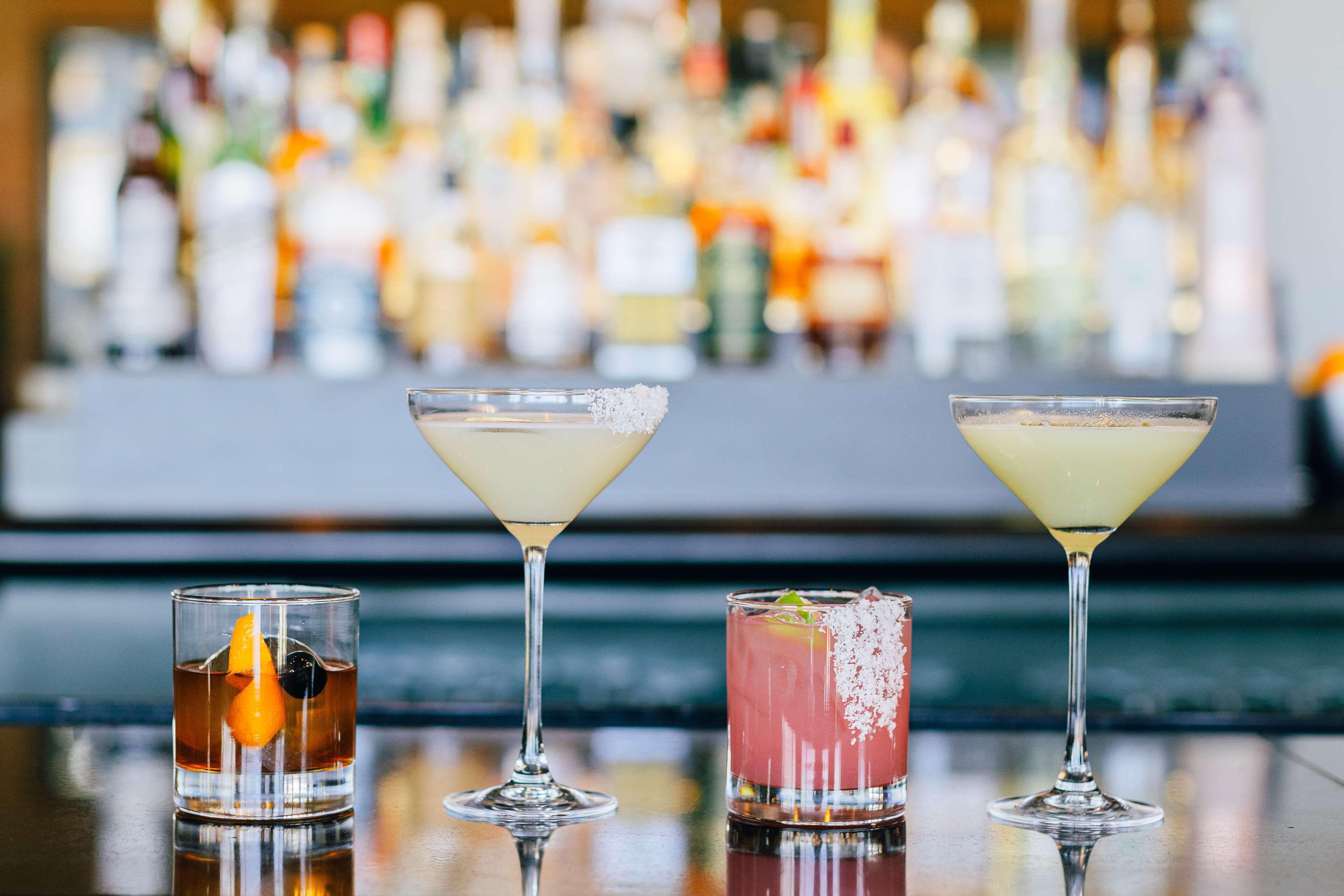 Three different cocktails on a bar counter with a blurred background of various bottles.
