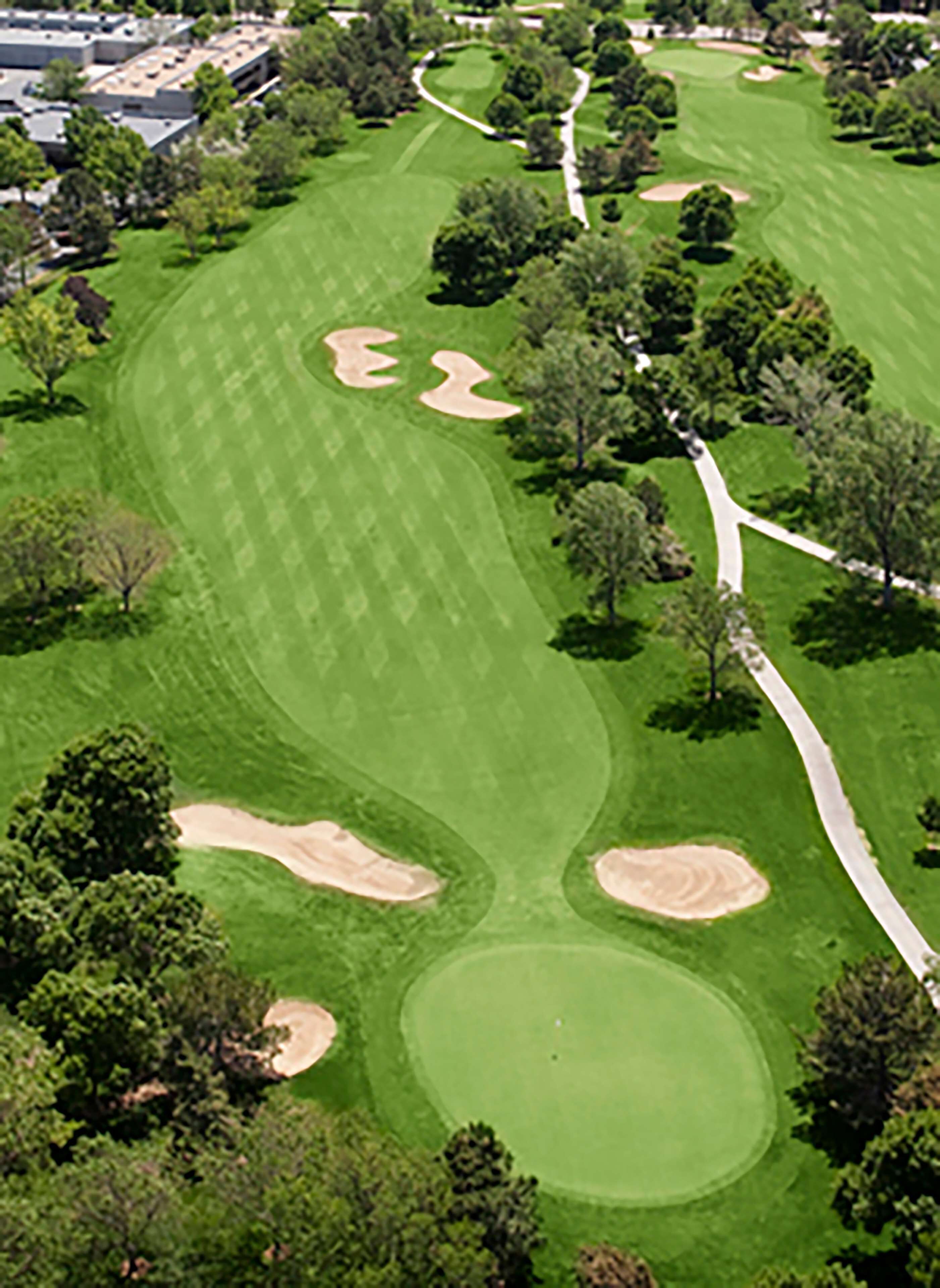 An aerial view of a golf course with sand bunkers, trees, and a clubhouse in the background.