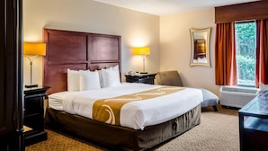 A hotel room with a large bed, two bedside tables with lamps, a chair, a desk, and a window with curtains. - Quality Inn Moss Point - Pascagoula (Moss Point)