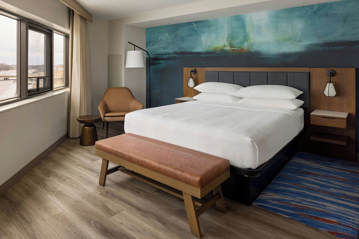 A hotel room with a large bed, a bench, a chair, a desk, and a painting on the wall.