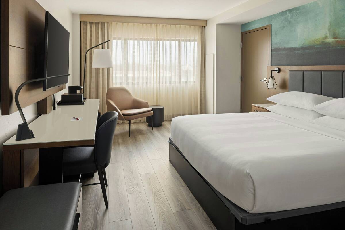 A hotel room with a bed, a desk with a chair, a TV, a lamp, and a window with curtains.