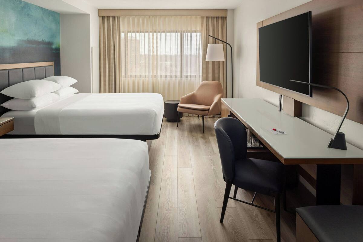 A hotel room with two beds, a desk, a chair, a TV, and a painting on the wall.