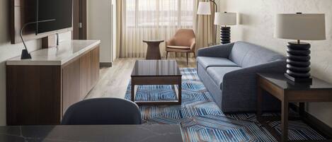 A hotel room with a flat-screen TV, a sofa, a coffee table, a desk, and a lamp.