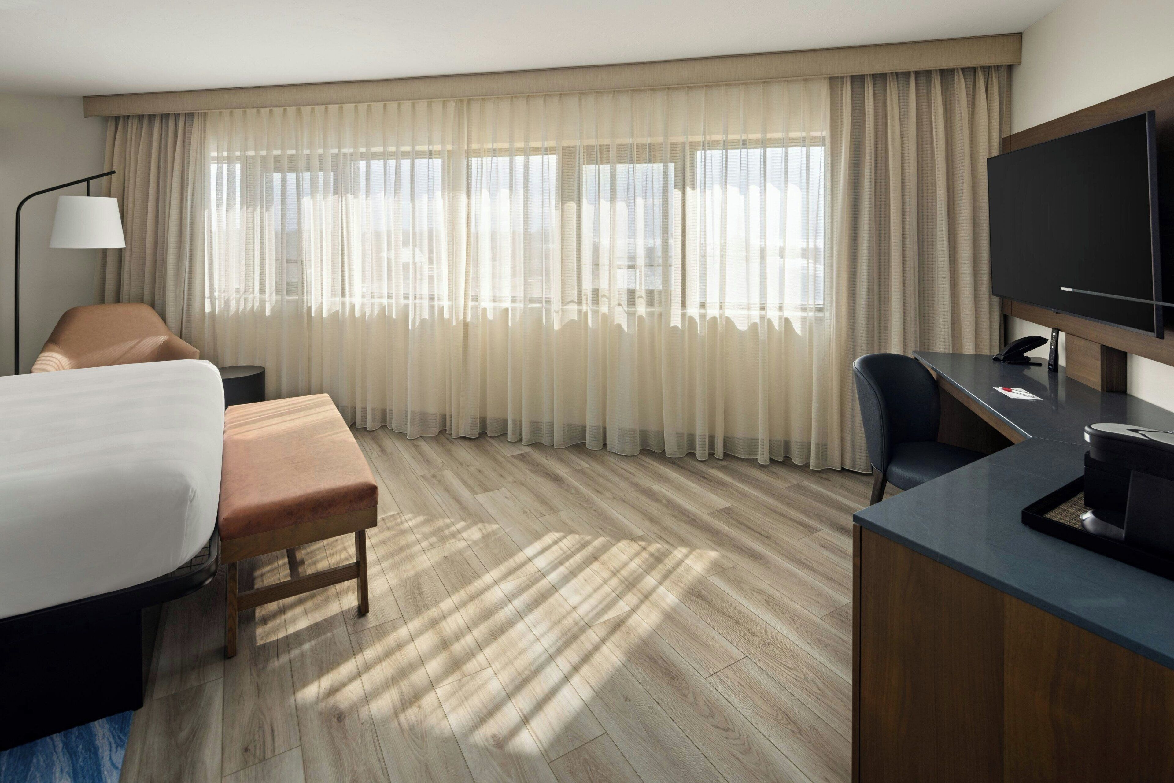 A hotel room with a bed, a desk, a chair, a TV, and a window with curtains.