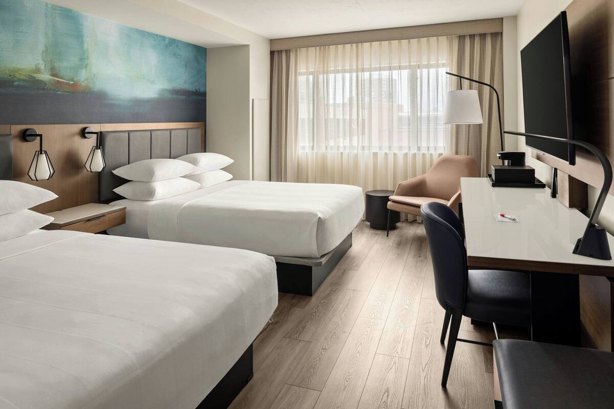 A hotel room with two beds, a desk, a chair, a TV, and a painting on the wall.
