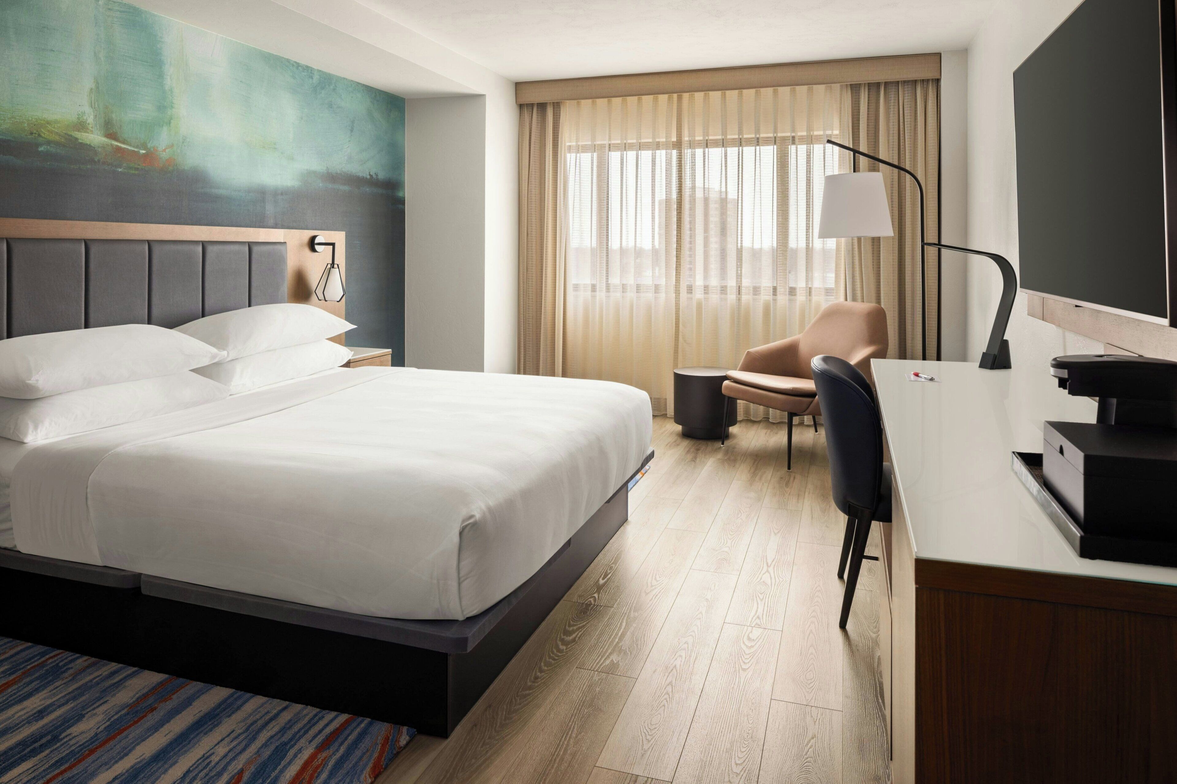 A hotel room with a large bed, a desk, a chair, a lamp, and a TV.