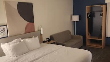 A hotel room with a large bed, a sofa, a desk, a coat rack, and a wall with a painting.