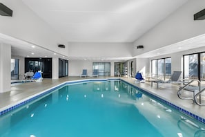 An indoor swimming pool with clear blue water, surrounded by lounge chairs and a tiled edge.