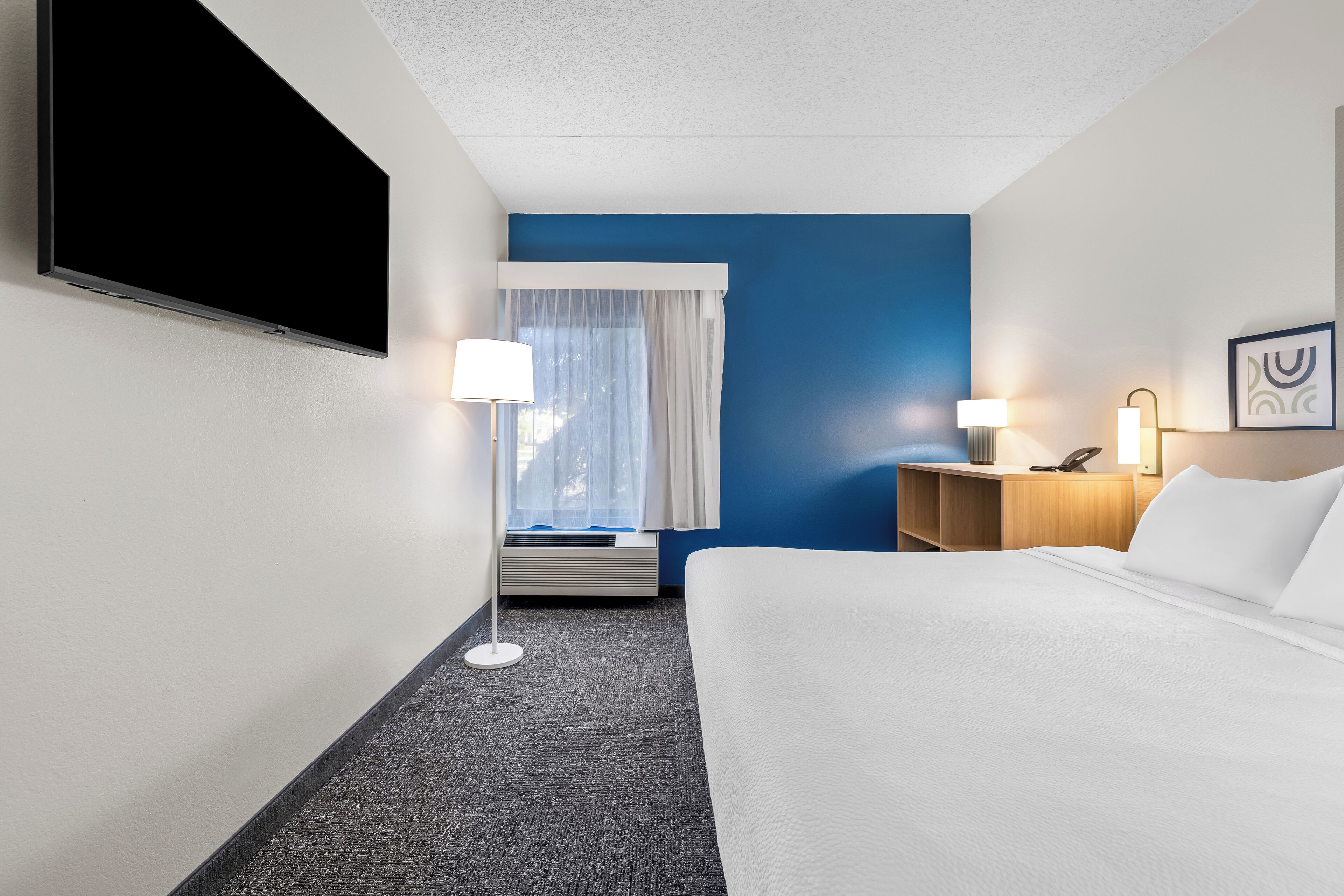 Habitación de hotel con cama, escritorio, silla, lámpara, televisor de pantalla plana y ventana con cortinas.