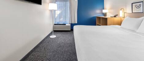 Habitación de hotel con cama, escritorio, silla, lámpara, televisor de pantalla plana y ventana con cortinas.