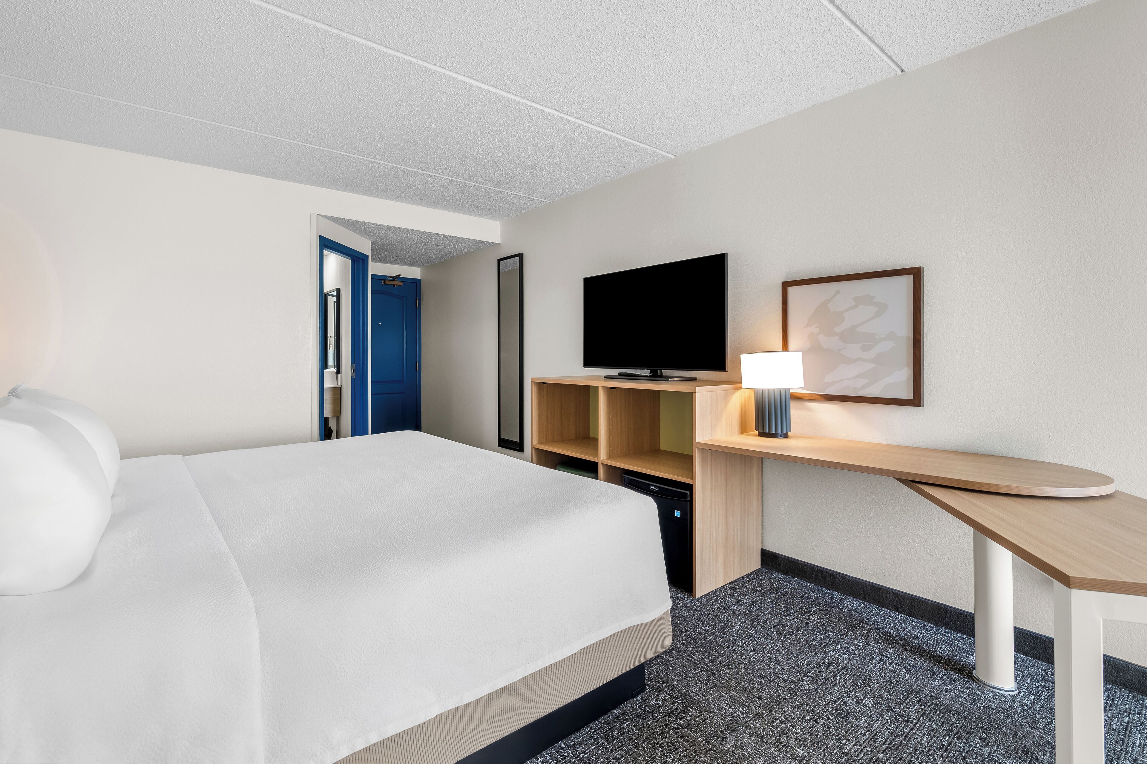 A hotel room with a bed, a television, a desk with a lamp, and a mirror on the wall.