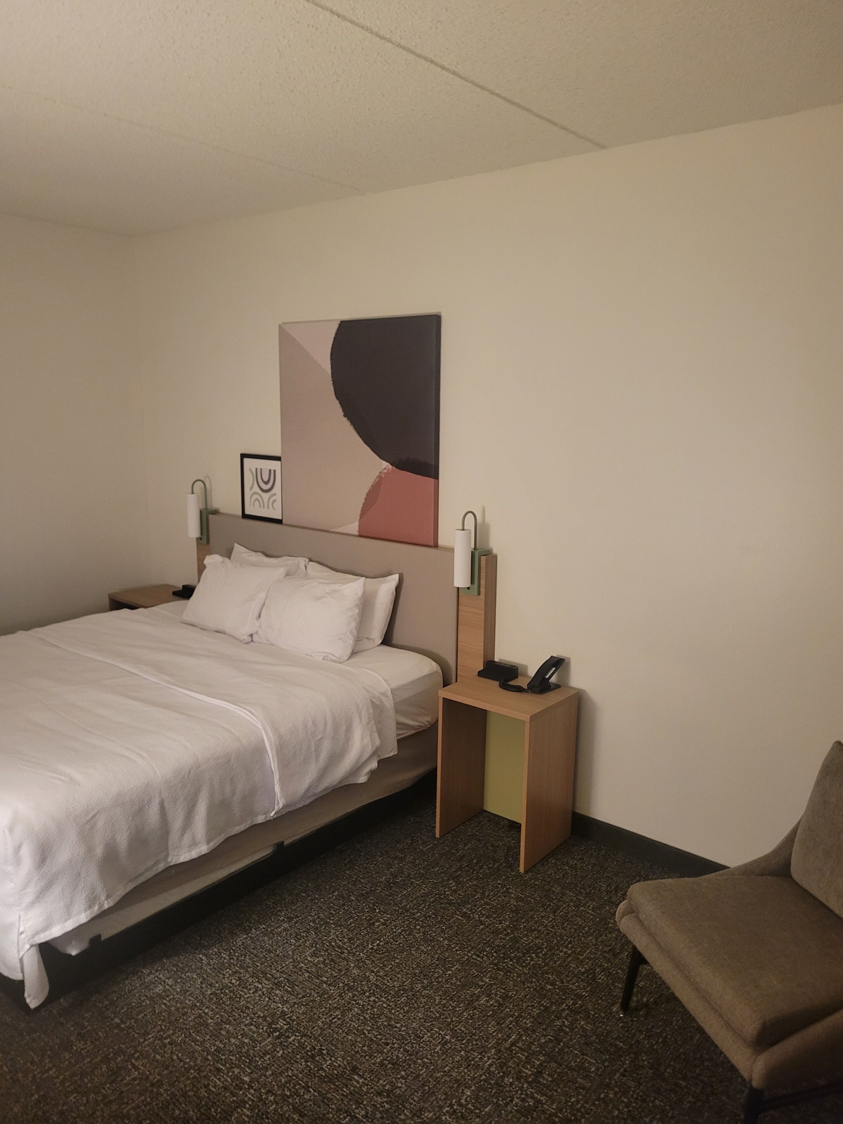 A hotel room with a bed, a nightstand with a phone, a chair, and a wall-mounted artwork.