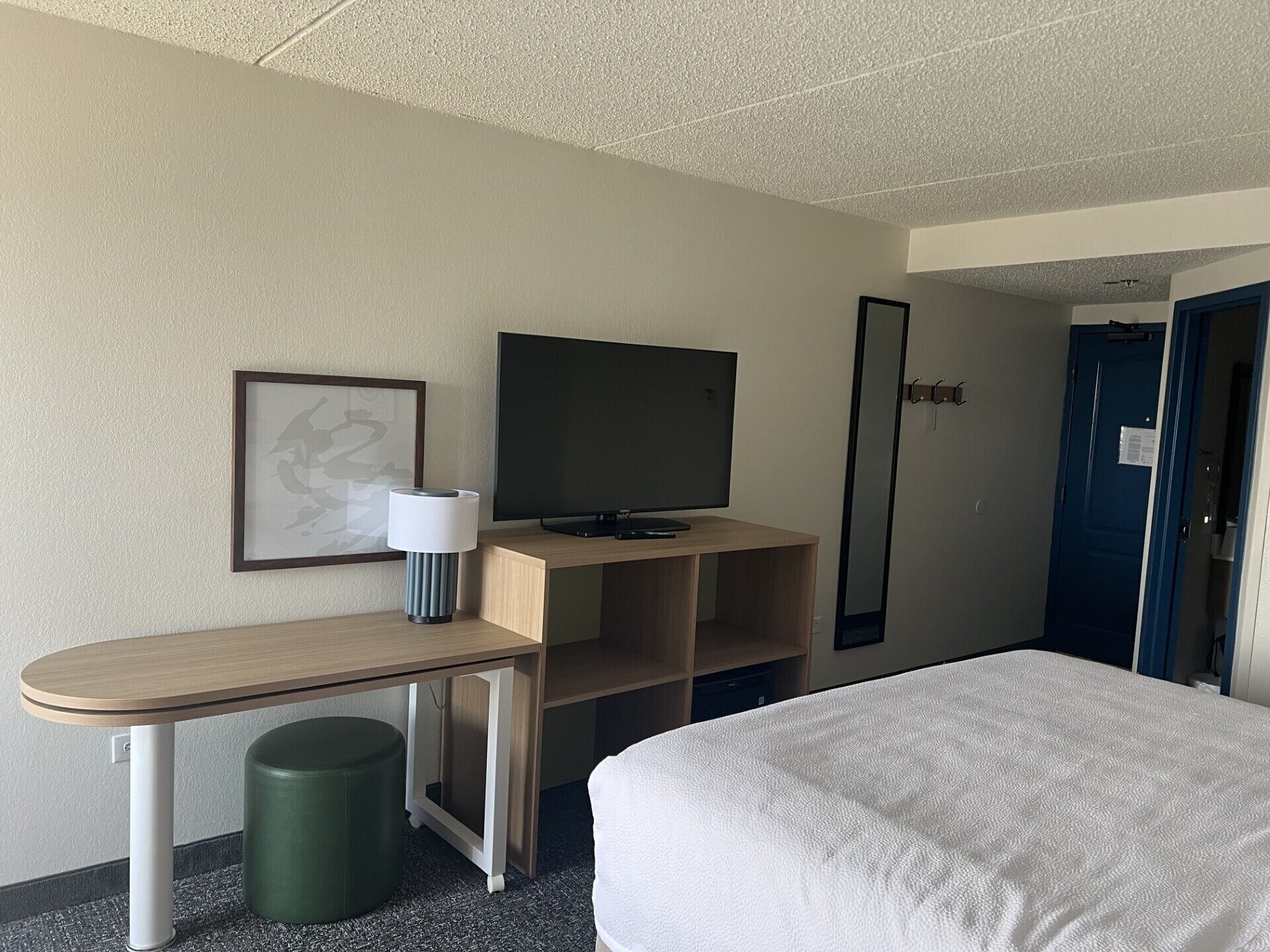 A hotel room with a bed, a desk with a lamp, a television, and a door.