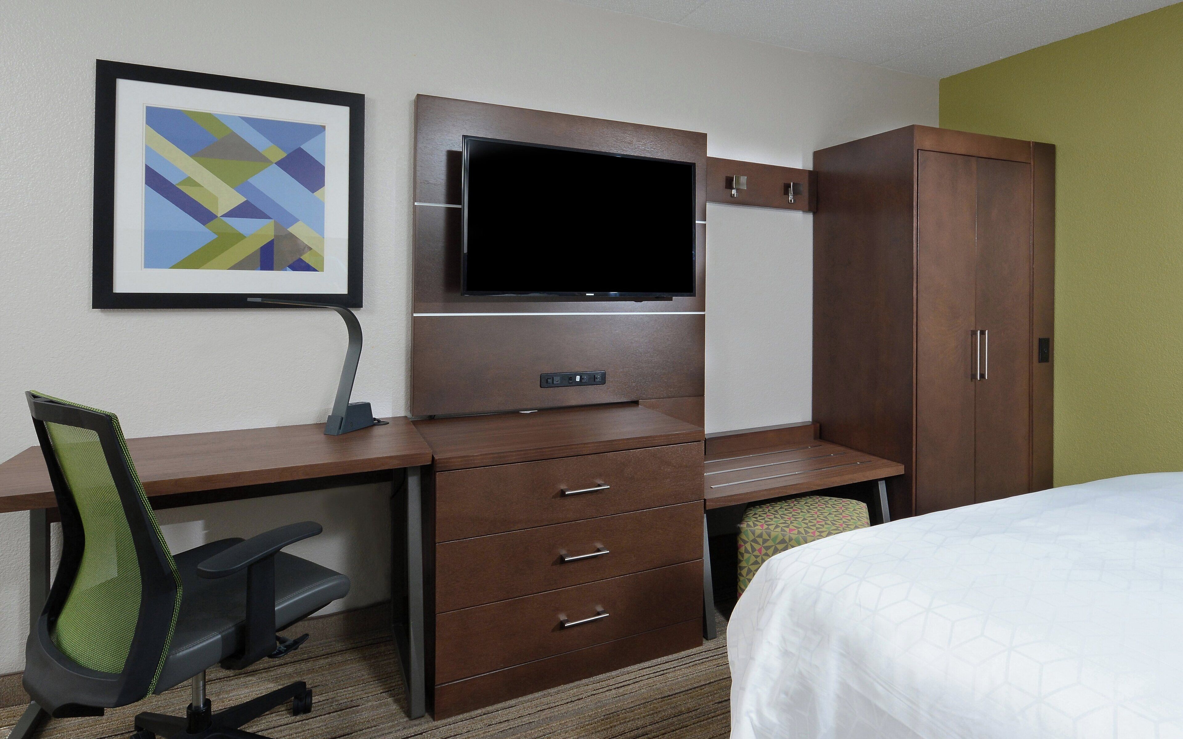 A hotel room with a bed, a desk with a chair, a TV, a wardrobe, and a framed picture on the wall.
