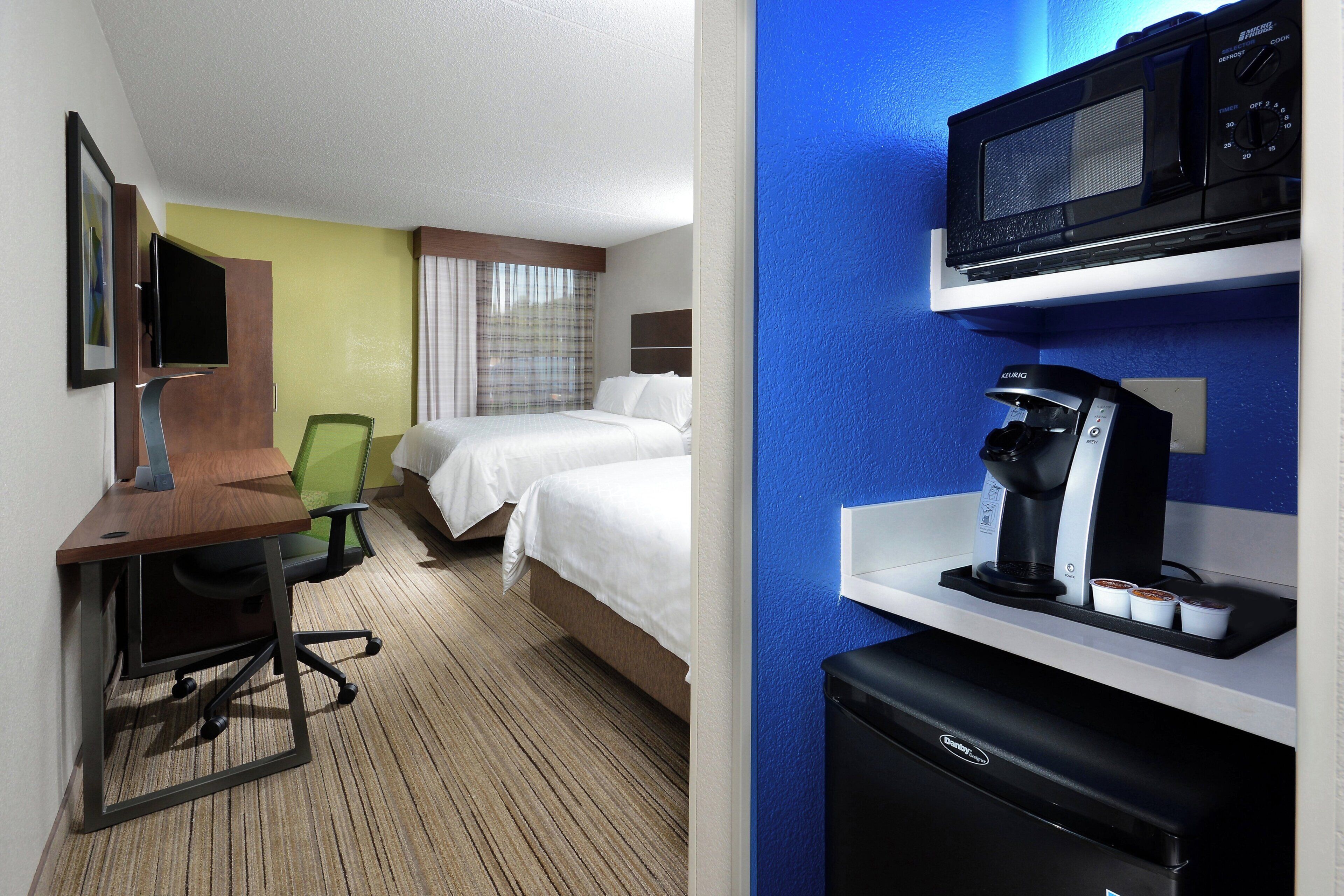 A hotel room with a desk, chair, bed, and a minibar with a coffee machine.