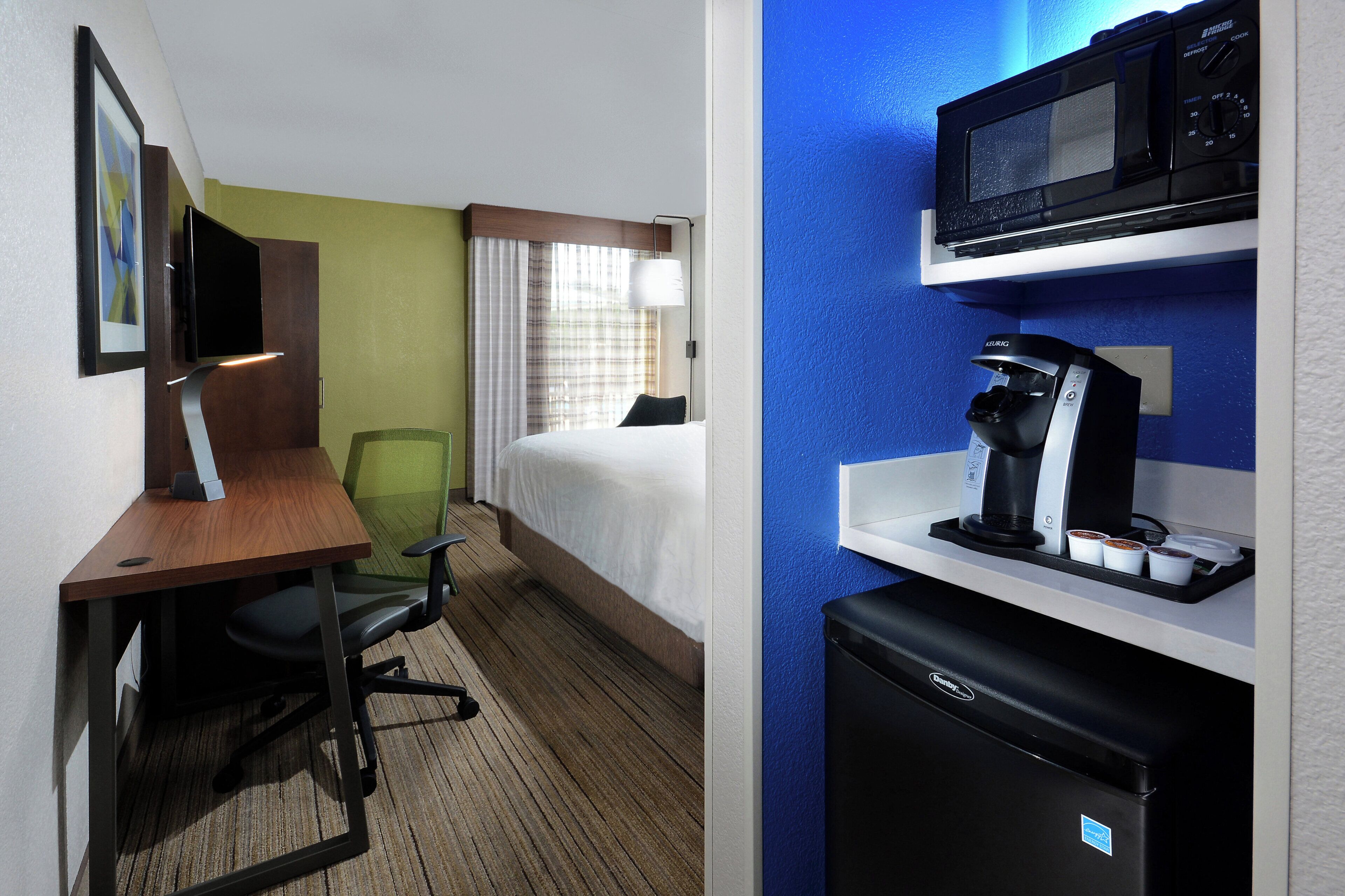 Habitación de hotel con cama, escritorio y silla, kitchenette con cafetera y televisión.