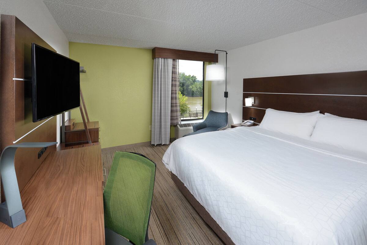 A hotel room with a large bed, a flat-screen TV mounted on the wall, a desk with a chair, and a window with curtains.