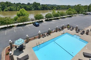 Une piscine entourée de chaises longues et d’une table, le tout bordé d’un parking et d’une rivière.