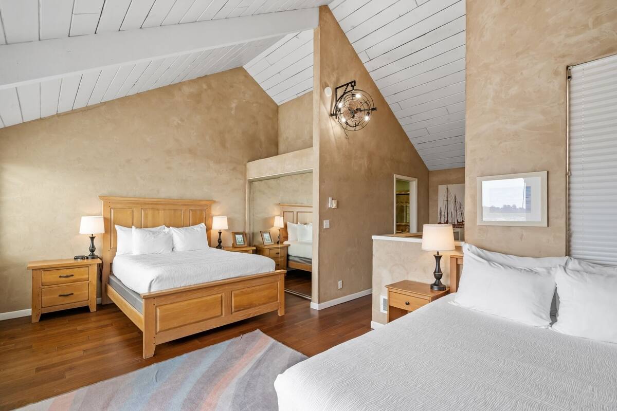 Un dormitorio con una cama grande, una cama más pequeña, mesitas de noche de madera, lámparas, un ventilador de techo y un espejo grande.