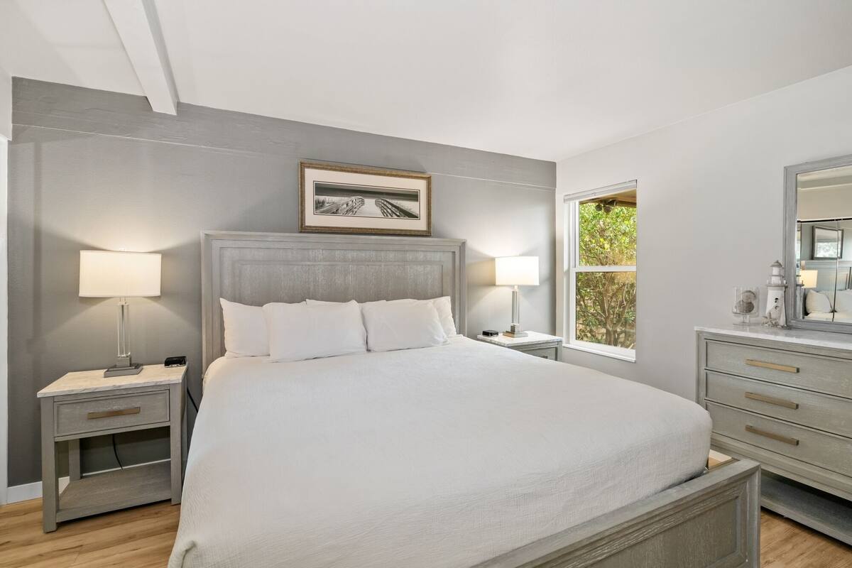 Un dormitorio con cama, dos mesitas de noche, un tocador, cómoda y ventana con vistas, además de un cuadro enmarcado sobre la cama.