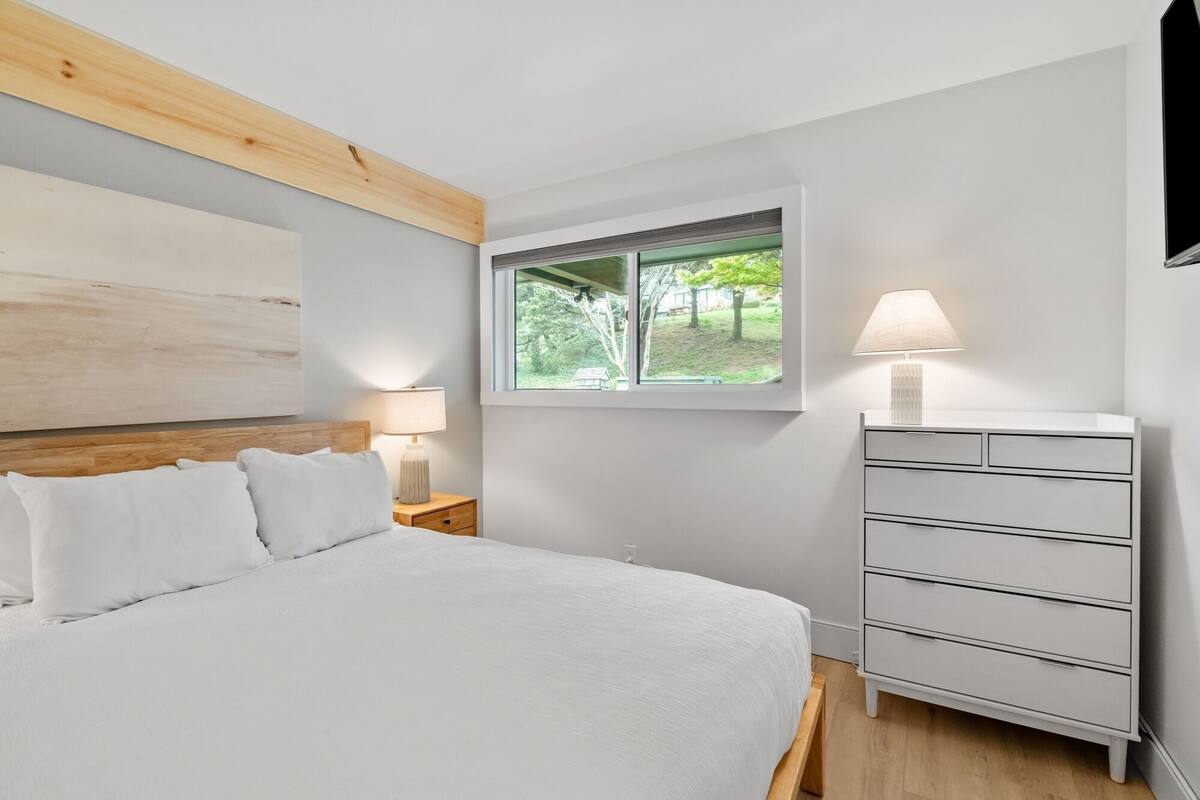 Un dormitorio con cama, mesita de noche, cómoda, ventana con vista a follaje y viga de techo de madera.