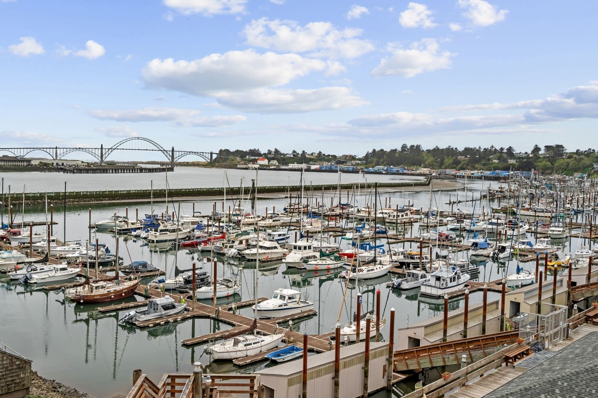 A marina with numerous boats docked, a bridge in the background, and a waterfront town.