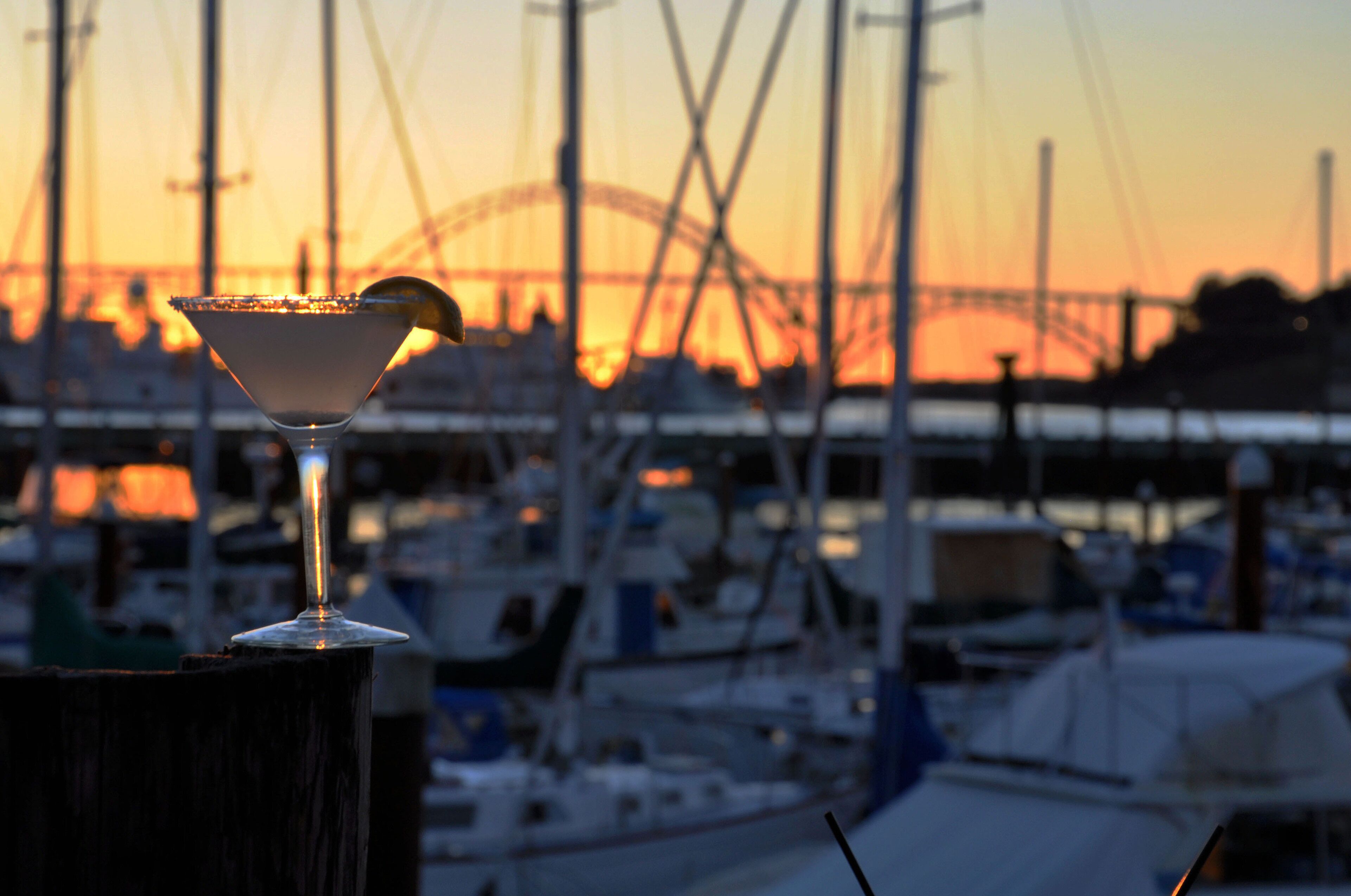 A martini glass with a lemon twist on a wooden surface, set against a marina backdrop with boats and a bridge during sunset.