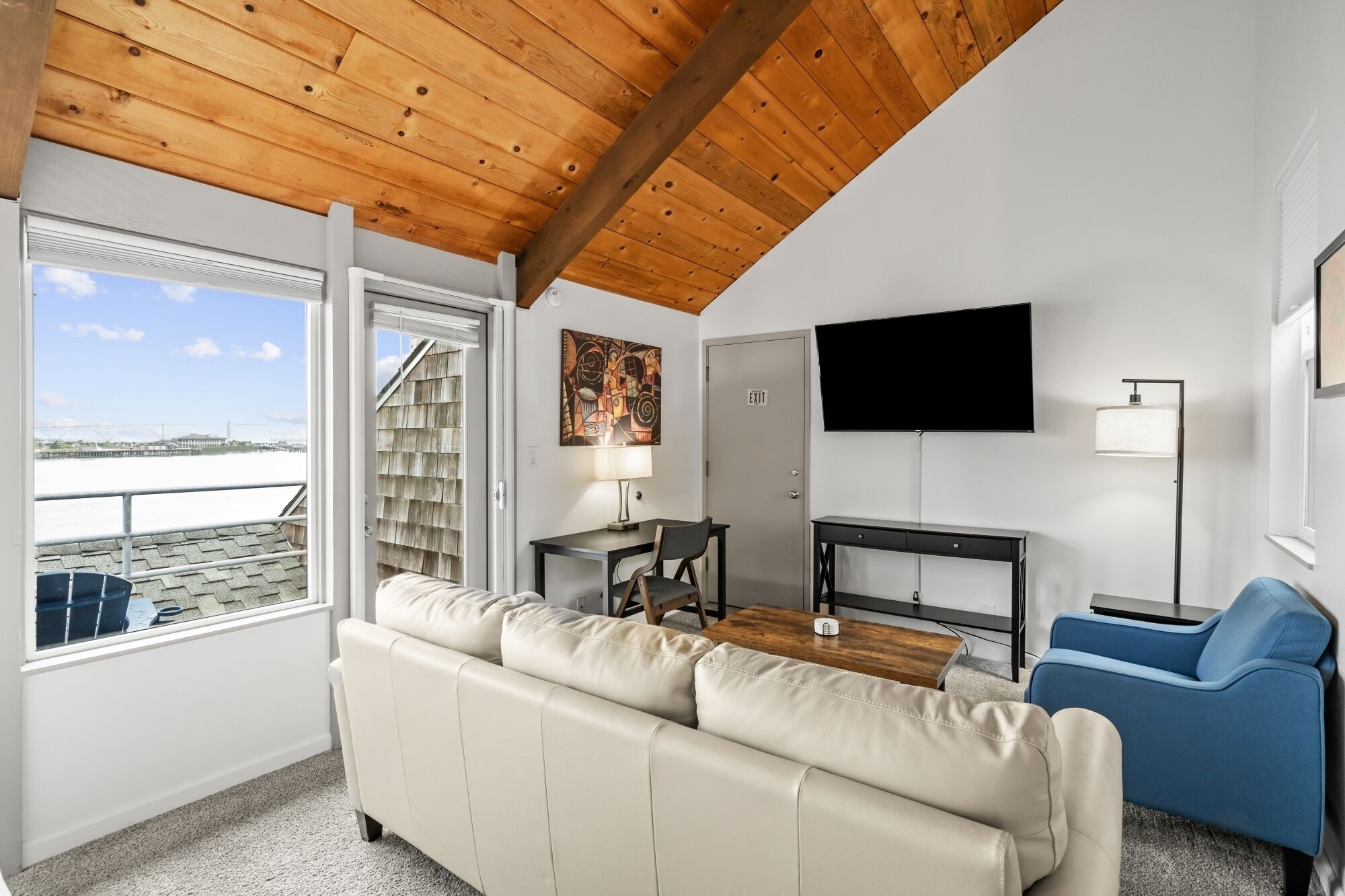 A room with a wooden ceiling, a sofa, a blue chair, a desk with a lamp, a television, and a view of the water outside.