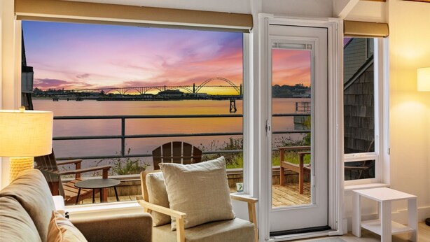 A room with a view of a sunset over a river, a sliding glass door, a sofa, a table, and a lamp. - Embarcadero Resort Hotel & Marina (Newport)